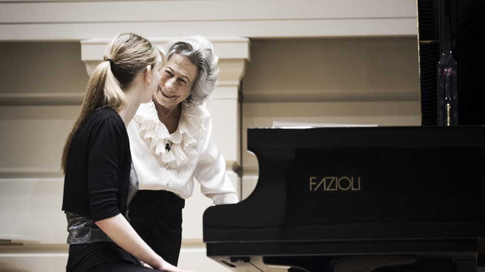 A women smiling and talking to a female student and the audience, standing next to a piano, with a female student wearing a black outfit, looking and listening, sitting at the piano.